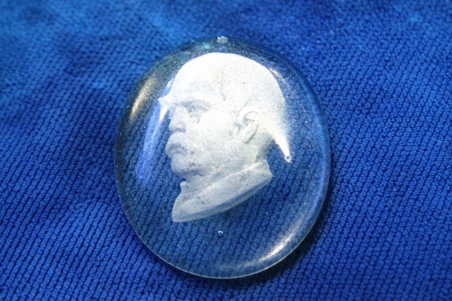 Oval glascame med profilbild av en man med mustasch på blå textil.